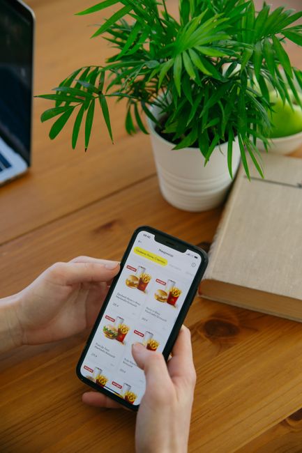 A close-up of a person's hand holding a smartphone displaying an online floral gift shop, with flower bouquet images visible on the screen. The scene includes a potted green palm plant with long, slen