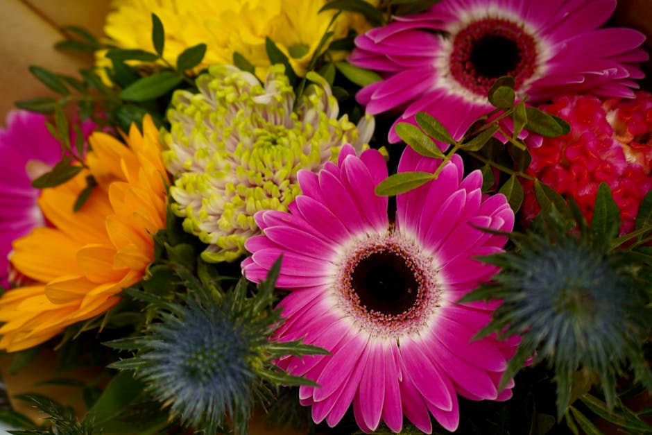 A vibrant floral arrangement featuring large pink gerbera daisies with dark centers, bright yellow daisies with orange accents, light green and white chrysanthemums, deep blue eryngium thistles with s