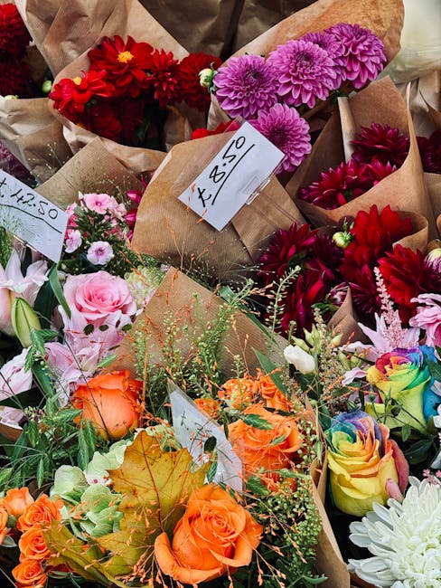 A vibrant assortment of fresh flower bouquets arranged on a display table, featuring red, pink, purple, orange, and multicolored roses, dahlias, and chrysanthemums. Each bouquet is wrapped in natural 