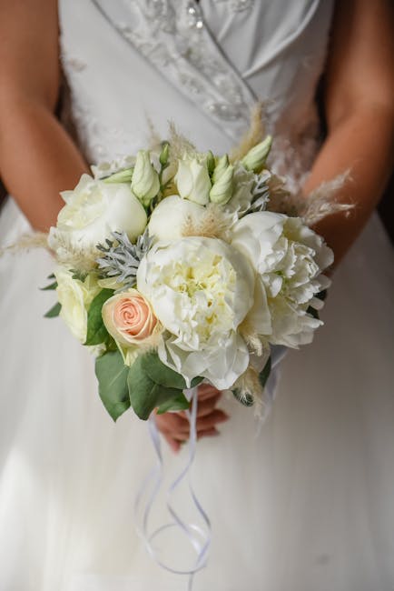 A bridal bouquet featuring a delicate arrangement of fresh white peonies, roses, and lisianthus, accented with soft grayish-green foliage and wisps of white feather-like accents. The flowers are tight