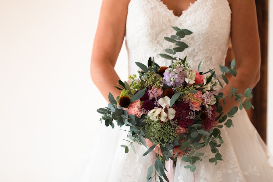 A bride holding a large bouquet arrangement of fresh flowers, including white, pink, purple, and burgundy blooms, accented with green foliage and eucalyptus leaves. The bouquet is wrapped with a soft 