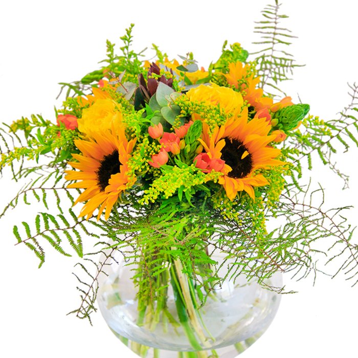 On first sight, this bouquet captures the mood of a joyful Colney Hatch morning, flush with sunshine and vibrant with colour. Grand sunflowers, their textured yellow petals unfurled, stand tall beside spirals of golden-yellow roses that exude a sense of warmth and invitation. The bouquet's abundant greenery, including trails of delicate, fresh fern fronds and shimmering eucalyptus, spills outwards, echoing the lushness found in hidden gardens off Colney Hatch Lane. Bright red Hypericum berries are tucked between blooms, their luminous orbs a playful nod to local parklands and adding a celebratory sparkle. Sprigs of light green goldenrod and slender green spikes complete the textured arrangement, their fine details reminiscent of the wild flora you might spot near New Southgate Cemetery. All these elements are anchored in a transparent, spherical glass vase, whose clear lines and water-filled base allow every vibrant stem and leaf to shine. The arrangement's overall scent is delicate yet uplifting-a fleeting memory of fresh fields and open skies typical of the season. Perfect for lifting spirits in a living room, as a centrepiece for dinner with friends, or as a gift to brighten a loved one's day, this bouquet expresses the unique charm of Colney Hatch through natural beauty and thoughtful design. Crafted with care by local hands, it invites a sense of celebration and comfort into any home.